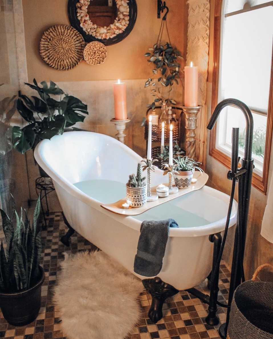 Banheiro estilo boho. Banheira branca com pés de metal, velas, plantas, vasos étnicos e tapete de cashmere. 