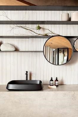 Banheiro minimalista. Bancada da pia em tom escuro do Topzstone Absulute Black, cuba preta, prateleiras e espelho redondo.