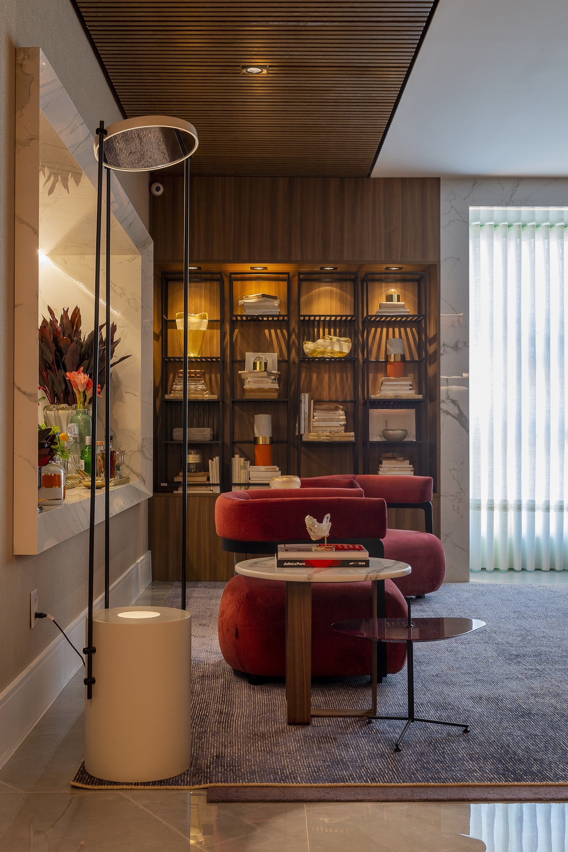 Casa Cor 2019: Linie Lounge Espaço com poltronas modernas vermelhas, parede do fundo revestida com madeiras e prateleiras, tapete com linhas e um abajur branco moderno.