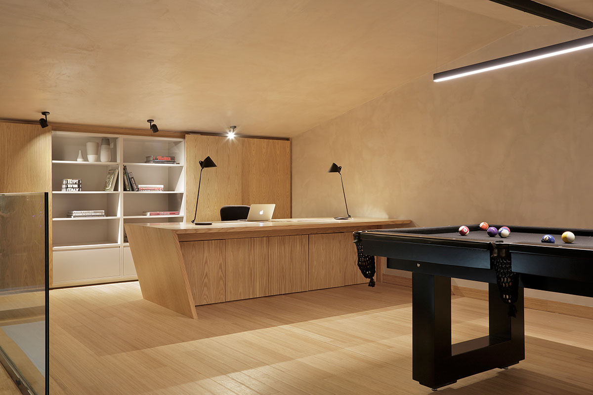 Ambiente com revestimento de madeira no piso, estante e mesa. Luminárias no teto e mesa do escritório. mesa de sinuca. 