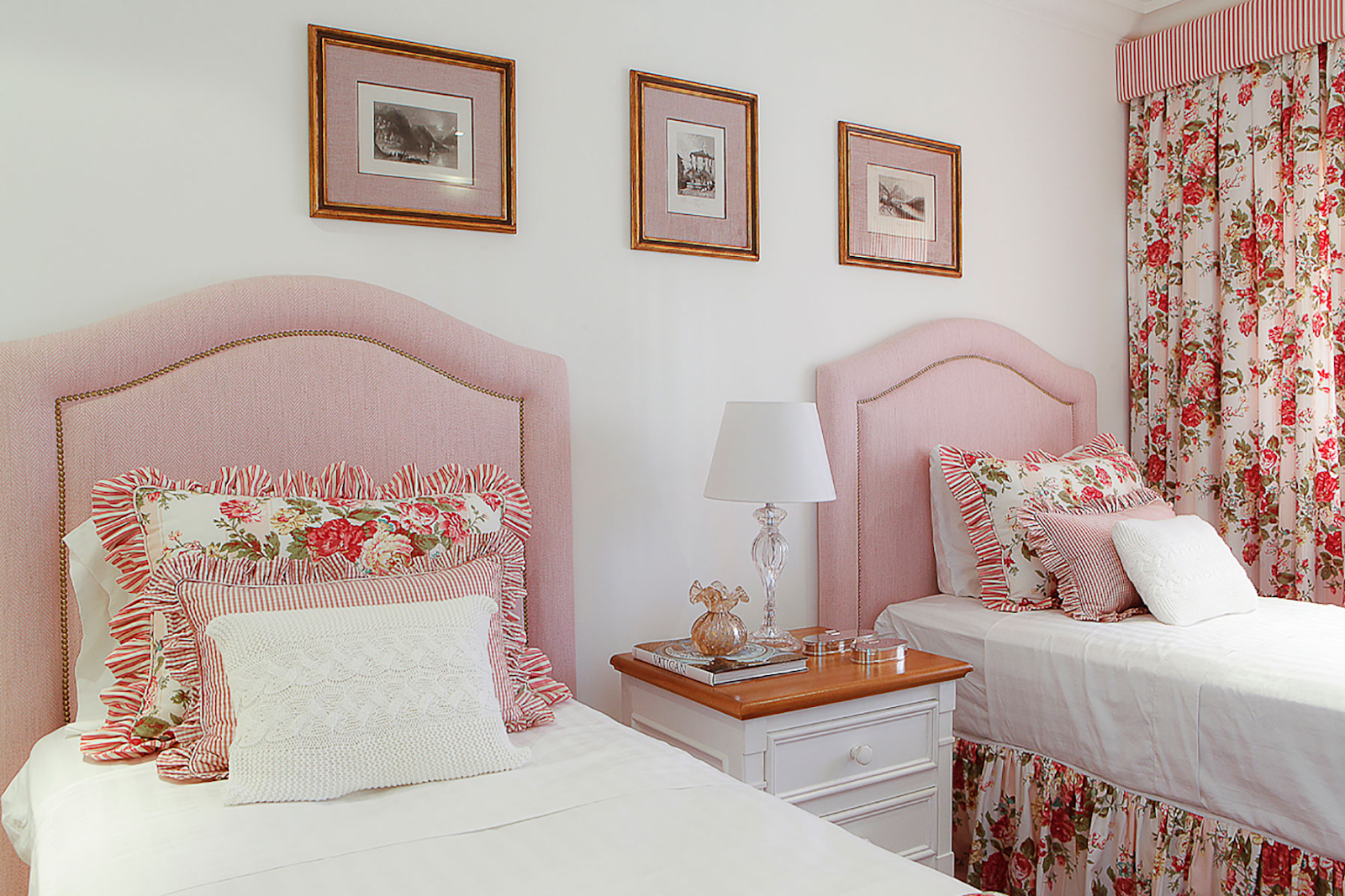 Quarto com cabeceira de cama rosa e cortina floral. Quadros na parede e criado mudo com marcenaria clássica.