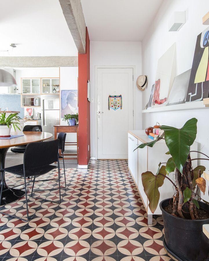 Hall de entrada de apartamento dividindo cozinha com ladrilhos azul e vermelho, parede pintada de branco, armários branco, mesa redonda e cadeiras pretas.