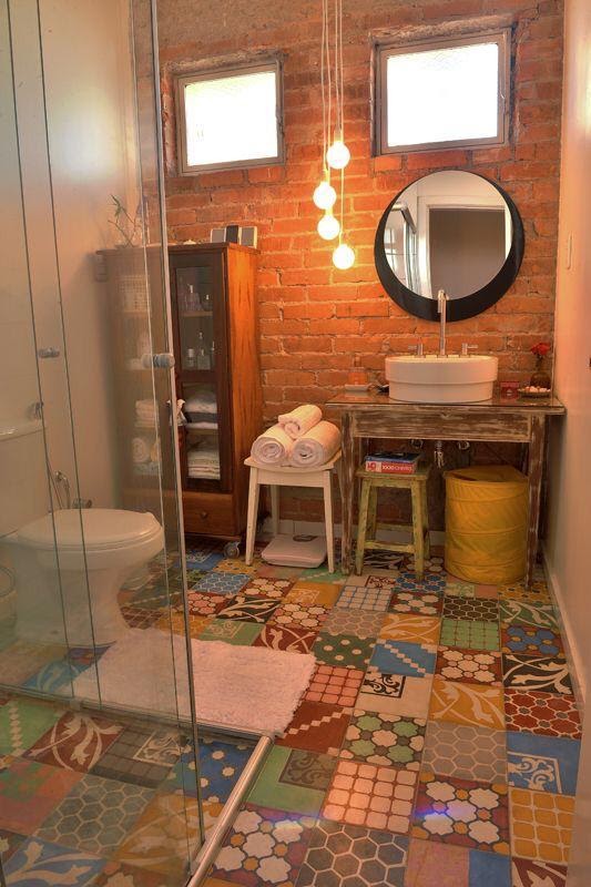 Banheiro com piso de ladrilhos coloridos, parede de tijolinhos, lâmpadas pendentes, armário de madeira com utensílios e box de vidro.