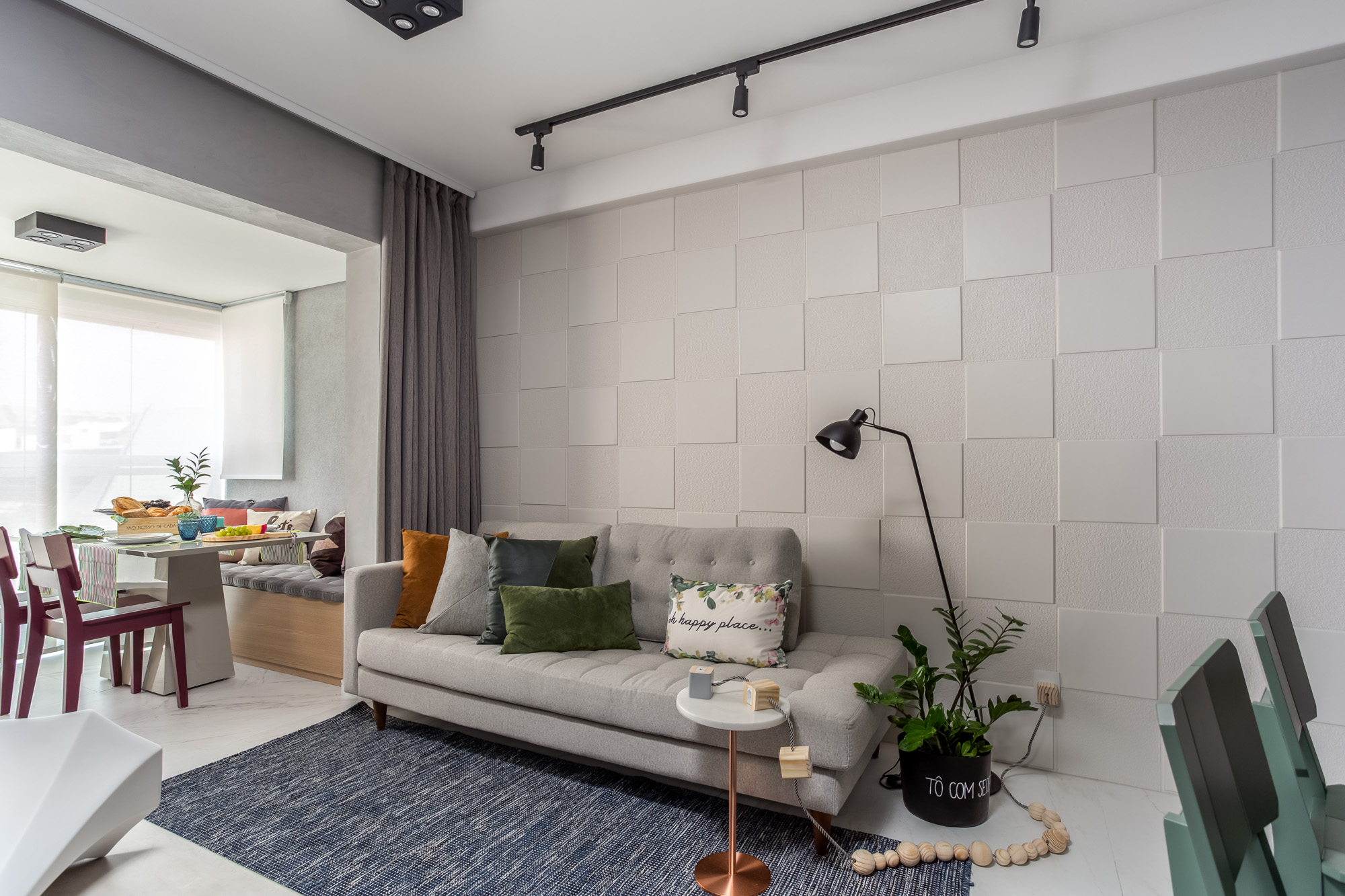 Sala de apartamento com revestimento cerâmico 3D em tons cinza, sofá cinza, almofadas coloridas e tapete azul compactado com ambiente de sala de jantar.
