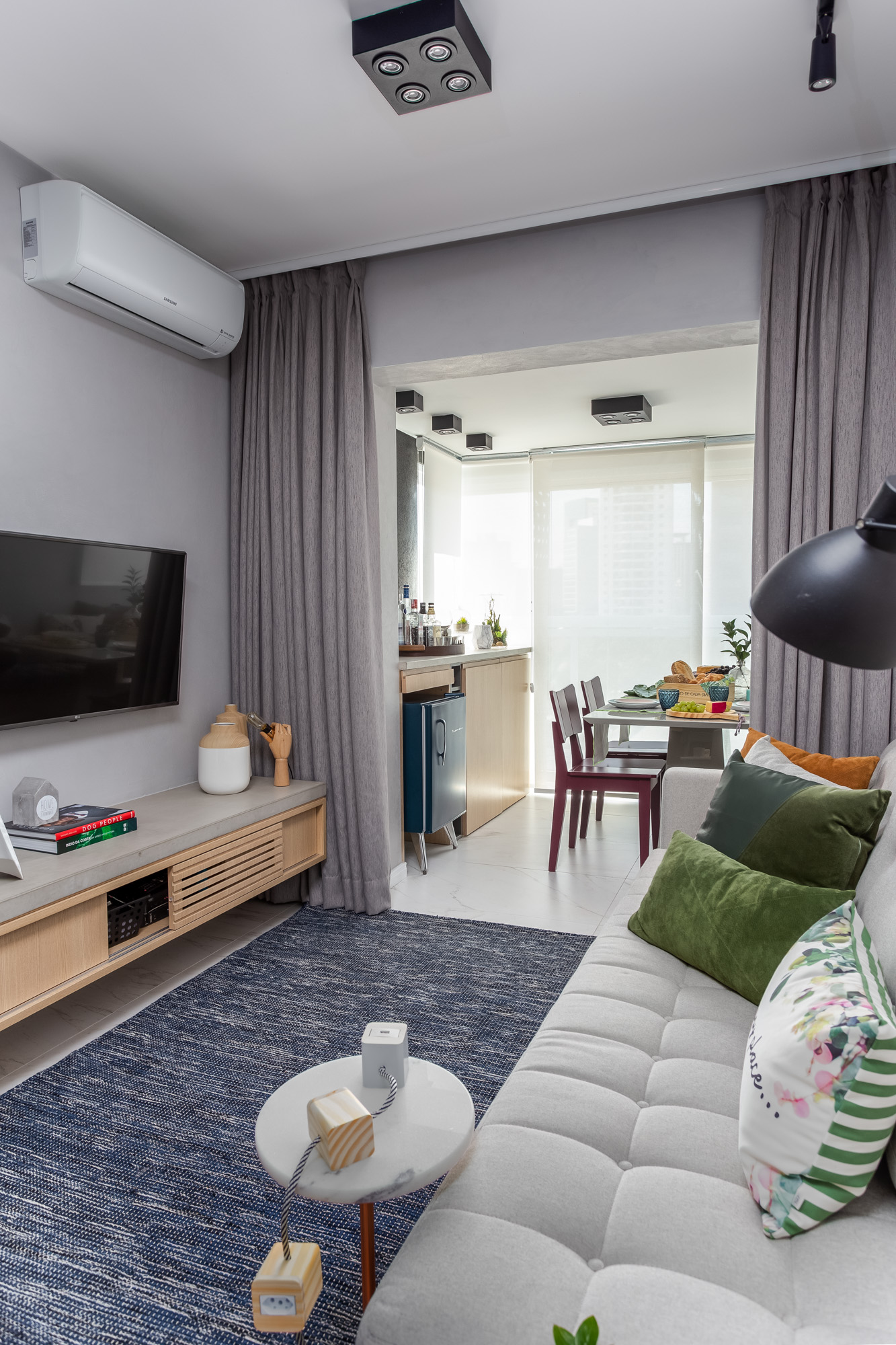 Sala de apartamento com sofá branco, almofadas coloridas, tapete azul e vista para sala de jantar