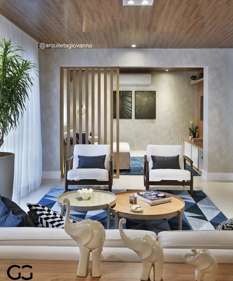 Sala de estar com poltronas e sofá brancos com almofadas e tapete em tons de azul, iluminação solar , teto com revestimento de madeira e decoração com plantas. 
