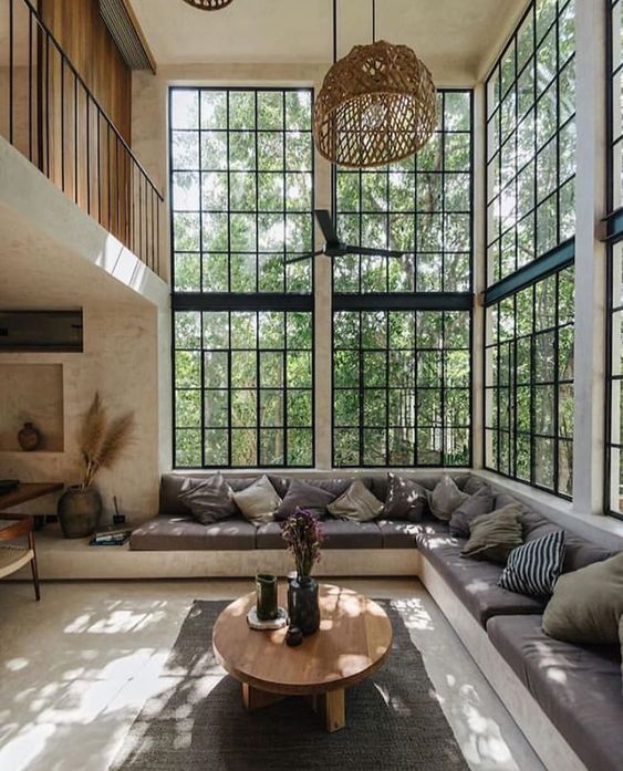 Sala de estar com sofá em L, almofadas cinzas, mesa de centro de madeira, ventilador de teto, janelas de vidro.