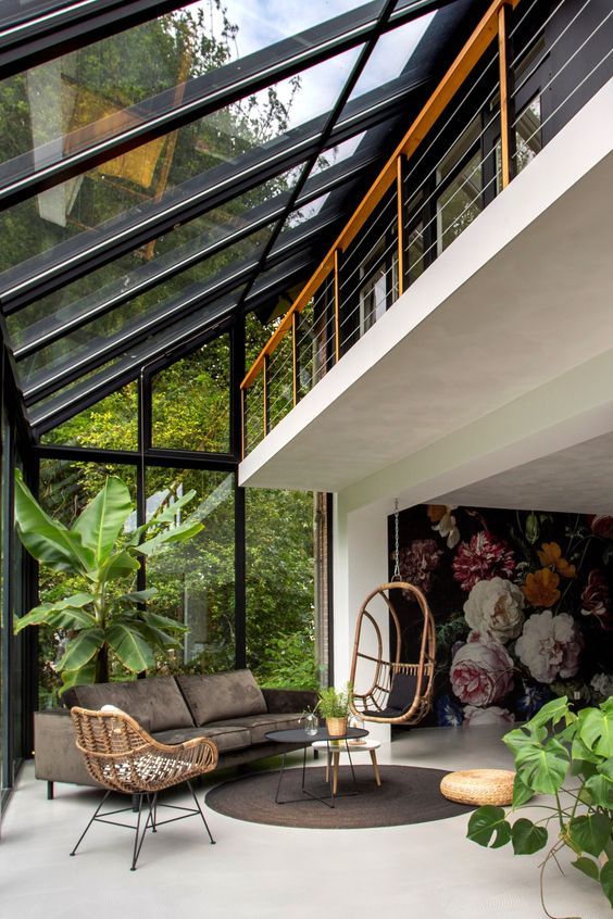 Ambiente envidraçado com cadeira de balanço, sofá grafite, plantas, mesinha de centro redonda e painel com flores coloridas na parede.