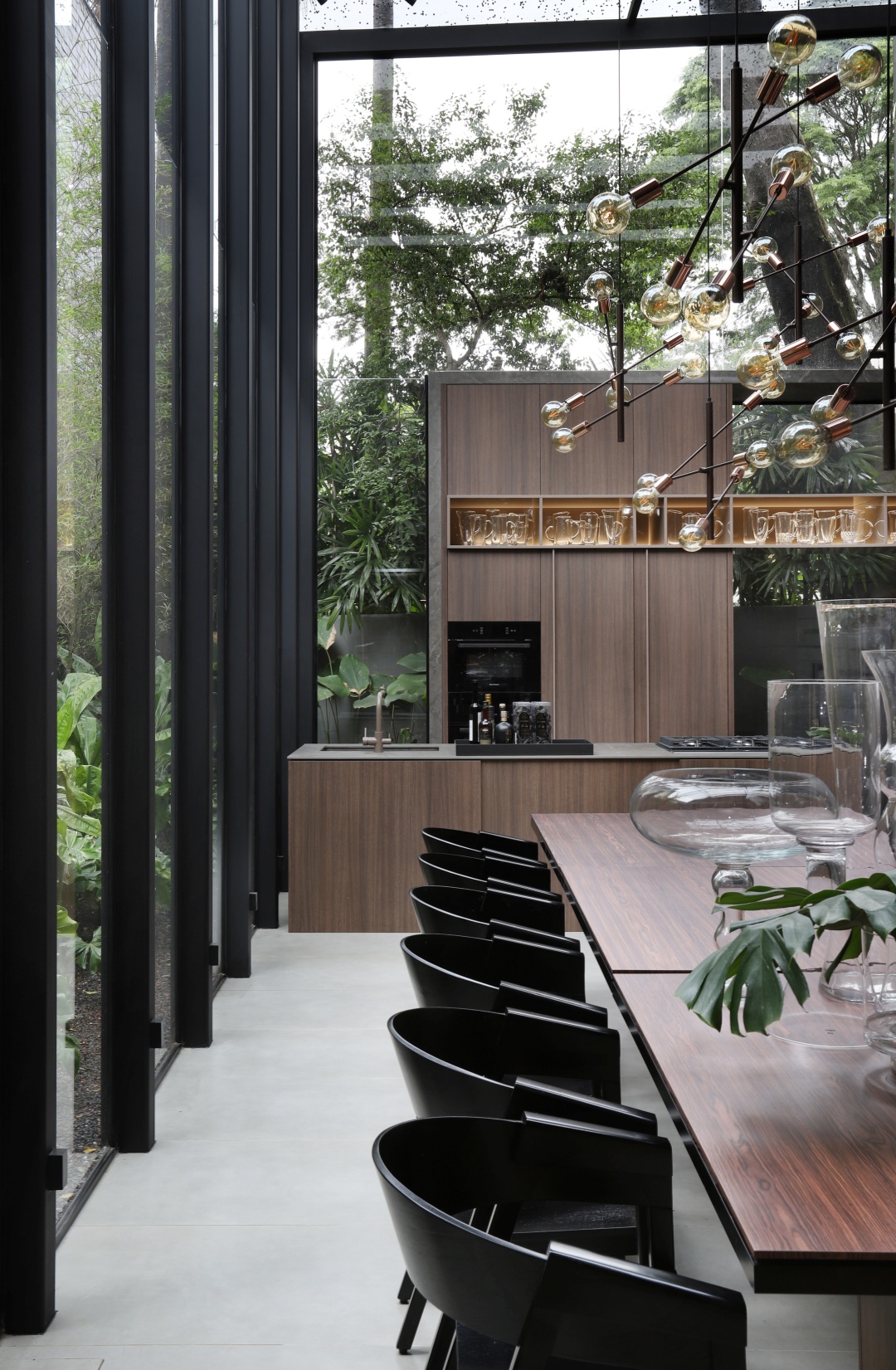 Cozinha gourmet com mesa grande de madeira com cadeiras pretas, vasos de vidro, luminária pendente, armário e bancada da cozinha amadeirados.