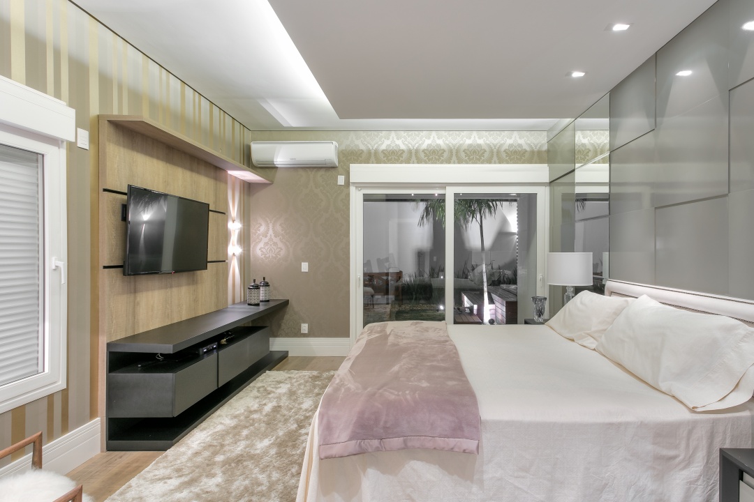 Dormitório em cores neutras com cama de casal, painel cinza na cabeceira da cama, bancada e painel de TV de madeira e iluminação para proporcionar maior conforto. 