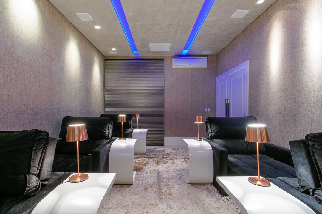 Sala de cinema com teto futurista e duas aberturas para que fossem embutidas LED em tom azul. Persianas pretas, luminárias na cor bronze e poltronas motorizadas em camurça preta..