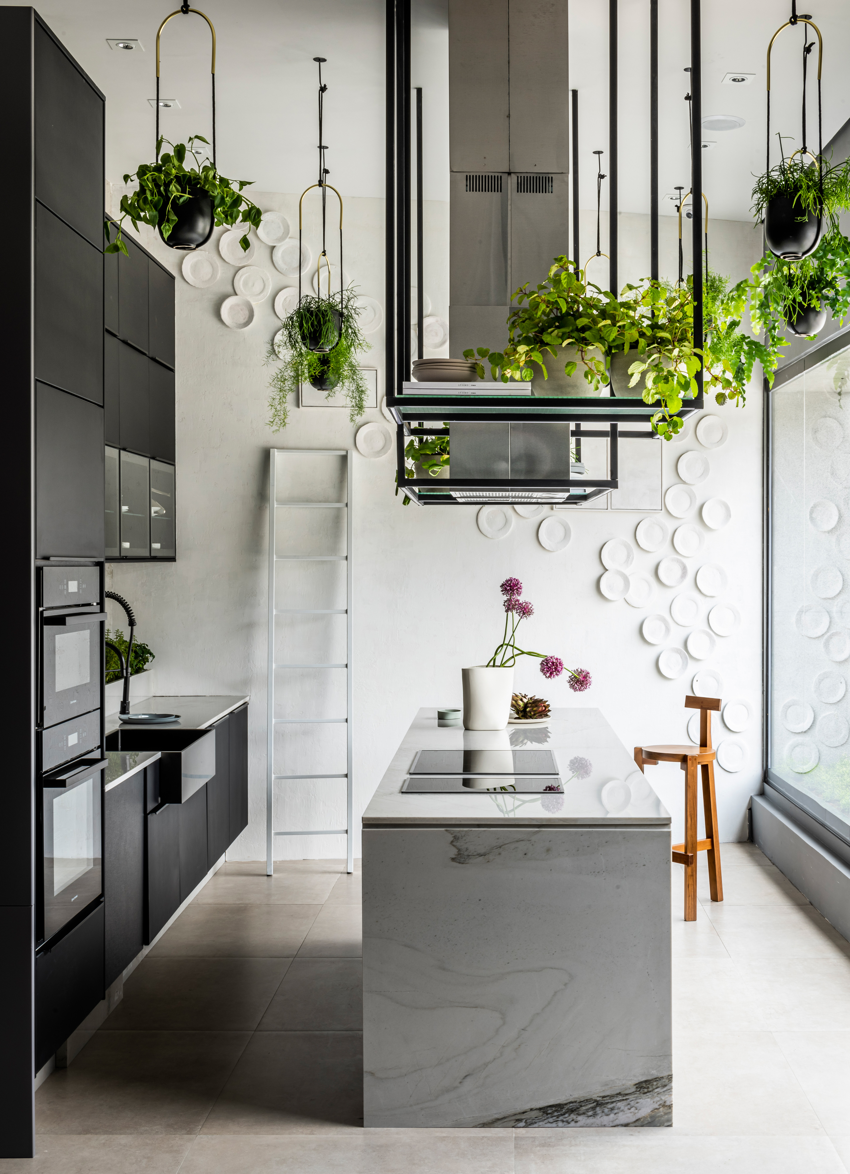 Cozinha com parede branca decorada com pratos brancos, armário de ferro de teto com muitos vasos de plantas, bancada de porcelanato com cooktop, armários preto e piso porcelanato neutro.