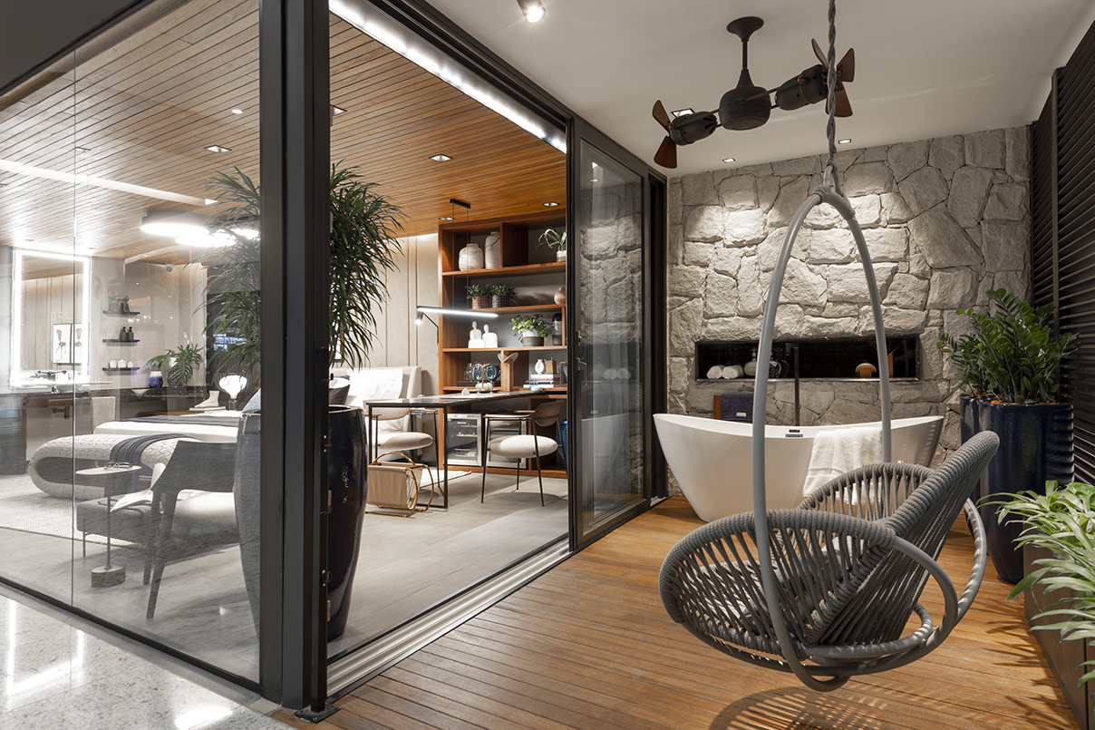 Janelas CASACOR  - Ambiente com piso de madeira, cadeira cinza de balanço, banheira branca, vista do ambiente interno com revestimento de madeira no teto, sofás brancos.