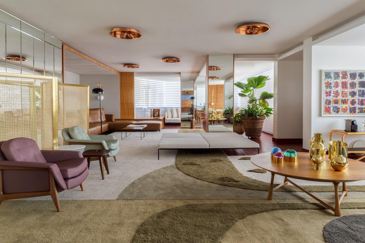 Sala de estar com poltronas d emadeira com estofado colorido, tapetes pelo chão, sofás branco, mesinhas de canto de madeira redonda, vasos de plantas, espelhos na parede.