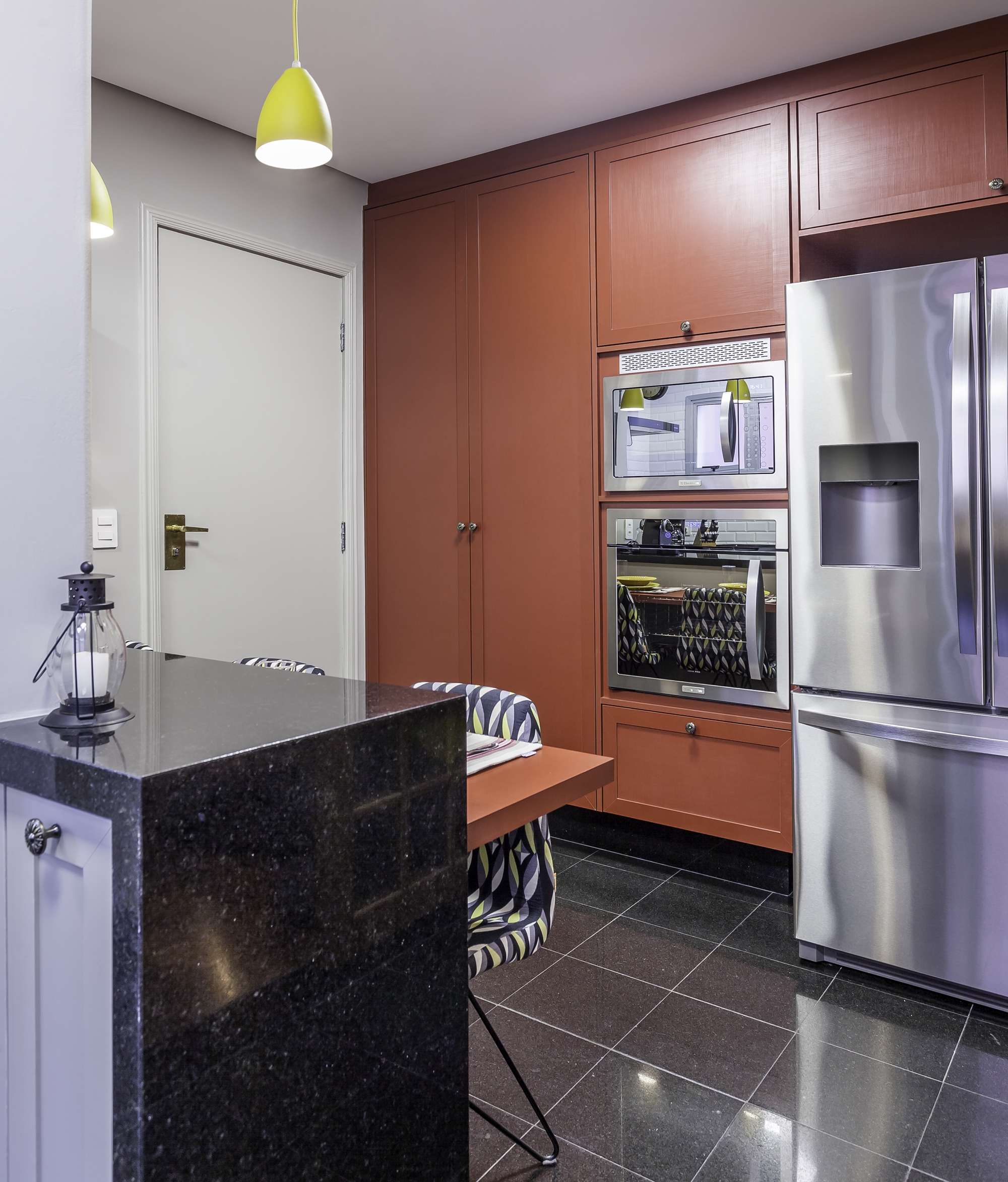 cozinha com decoração de personalidade, armários e mesa vermelhos, piso de granito preto, eletrodomésticos de inox, luminárias pendentes amarelas e bancada de mármore preto.