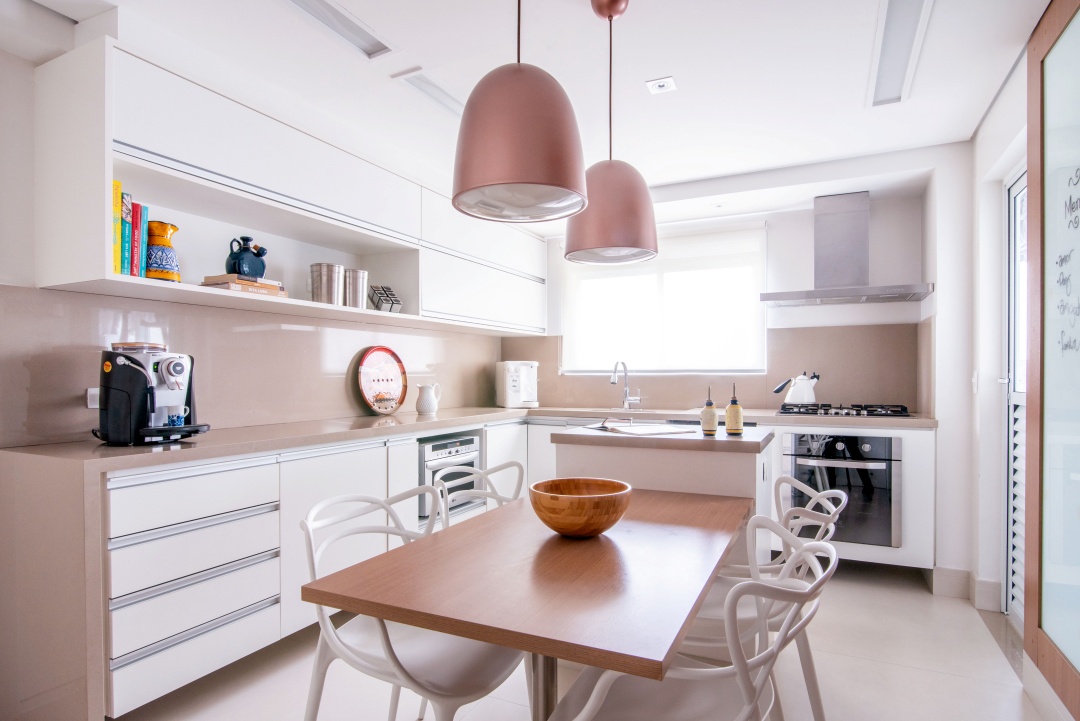 Cozinha com armários em tons neutros, piso em tom neutro, luminárias pendentes bronze e mesa de madeira com cadeiras brancas.