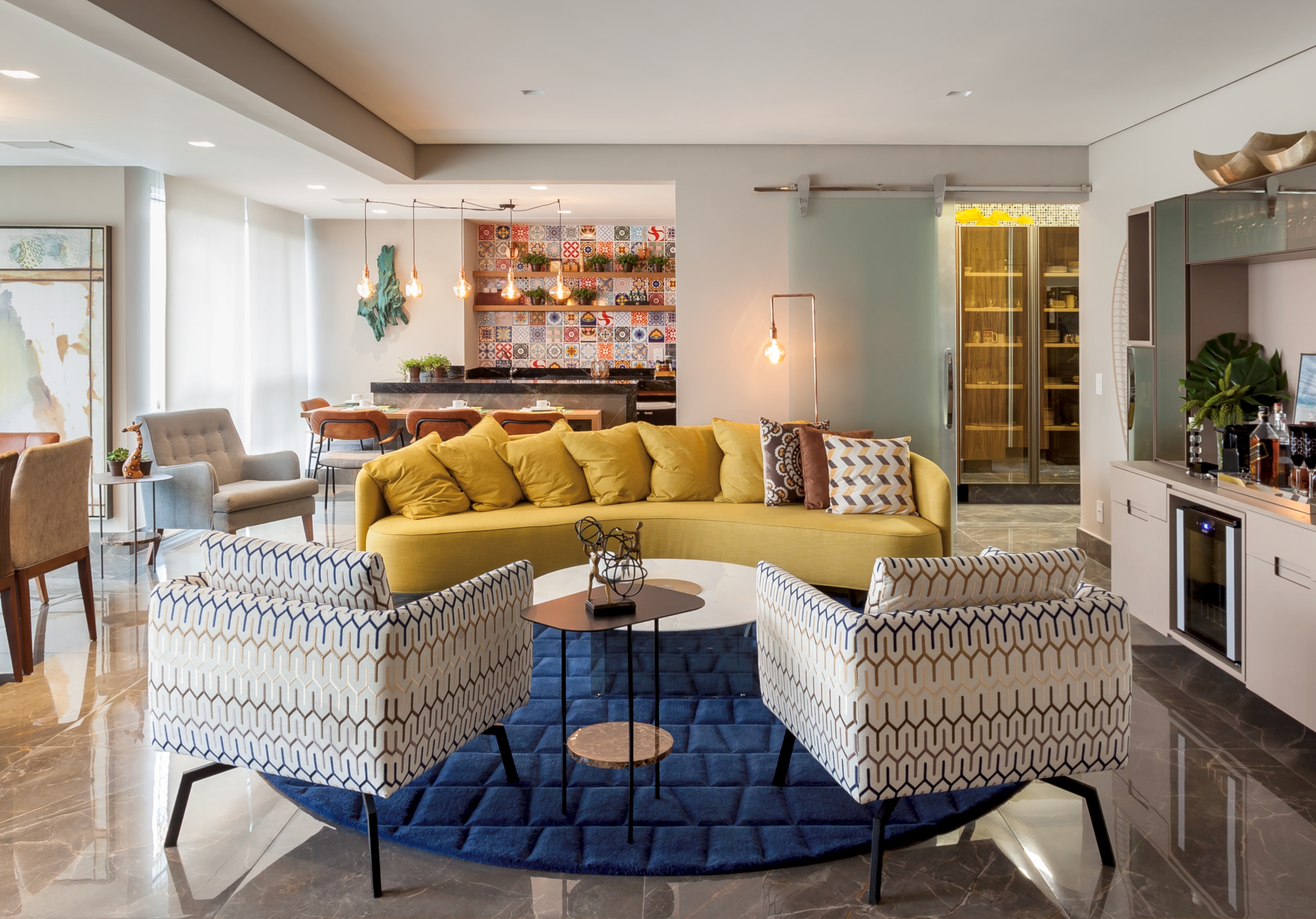 Sala com poltronas branca personalizada com desenhos geométricos coloridos, sofá curvo amarelo e tapete azul, paredes brancas e mesinha de centro redonda branca.