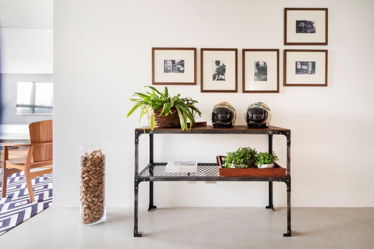 Aparador vintage de dois andares com vasinhos de plantas e dois capacetes apoiados, parede de fundo branca com quadros decorativos preto e branco.