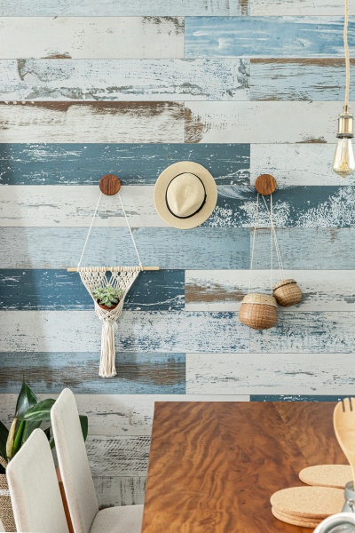 Decoração de casa de praia - Vista da mesa de jantar de madeira e parede de fundo com pintura imitando madeira em tons de azul com chapéu, e objetos de palha pendurados e espelho grande encostado na parede.