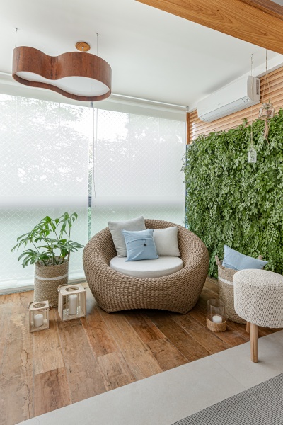  Sacada com jardim vertical na parede lateral, piso de madeira, sofá de palha com estofado branco e almofadas claras, bancos e cestos de palha e velas no chão.