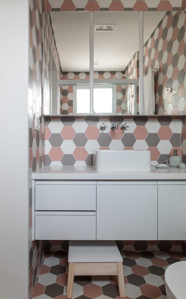 Revestimentos estampados: Banheiro com revestimento hexagonal nas paredes e piso em uma combinação de branco, cinza e rosa antigo, armários brancos e banquinho de madeira com estofado branco