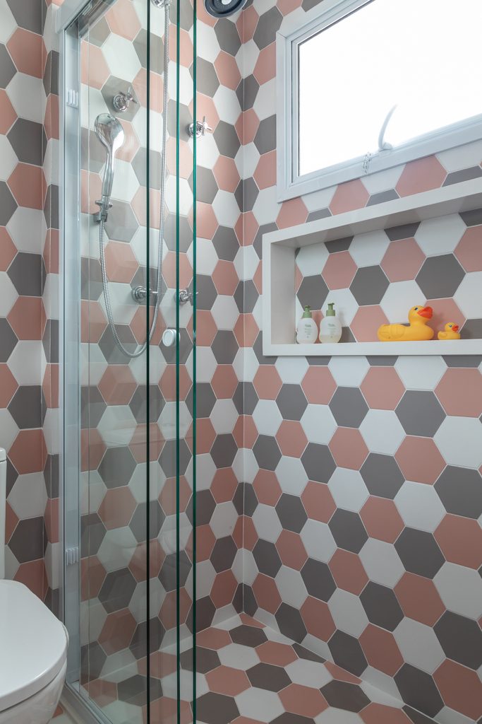 Revestimentos estampados: Banheiro com revestimento hexagonal nas paredes e piso em uma combinação de branco, cinza e rosa antigo, box de vidro e nicho branco.