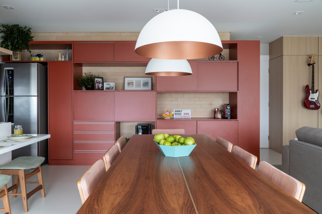 Cozinhas coloridas - cozinha com armário goiaba, mesa de madeira com cadeiras estofada rose, piso porcelanato branco e luminária pendente bronze.