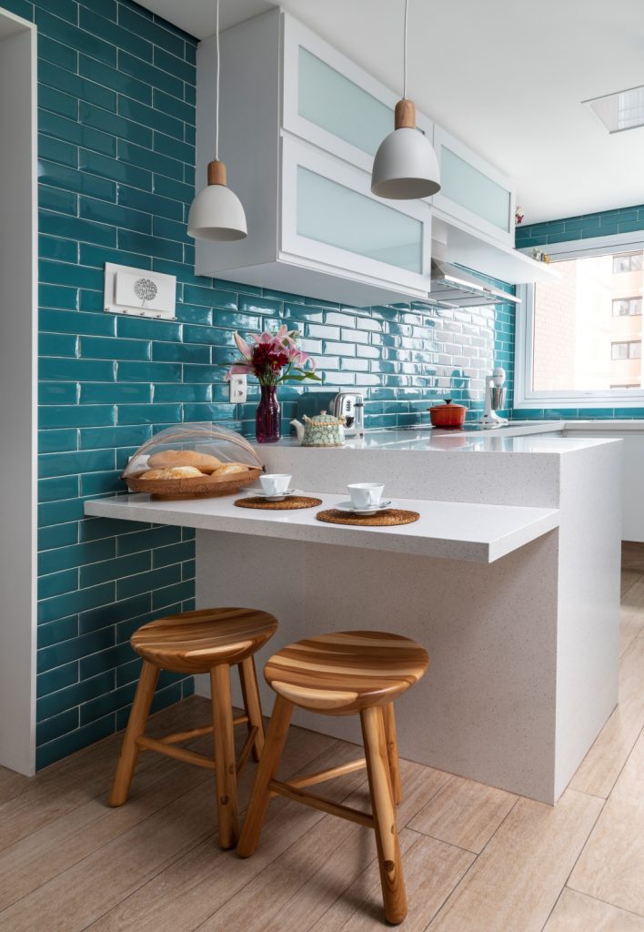Cozinhas coloridas - cozinha com azulejo subway tiles verde por toda parede, bancadas e armários brancos, piso de madeira, banquetas de madeira e luminárias pendentes brancas. 