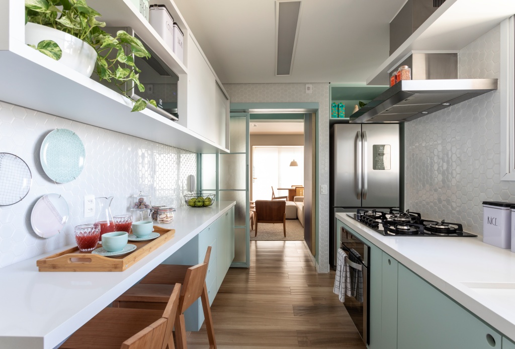 Cozinha com armários verde claro e branco, piso de madeira, cadeiras de madeira, acessórios verde claro e revestimento branco hexagonal em formato pequeno nas paredes.
