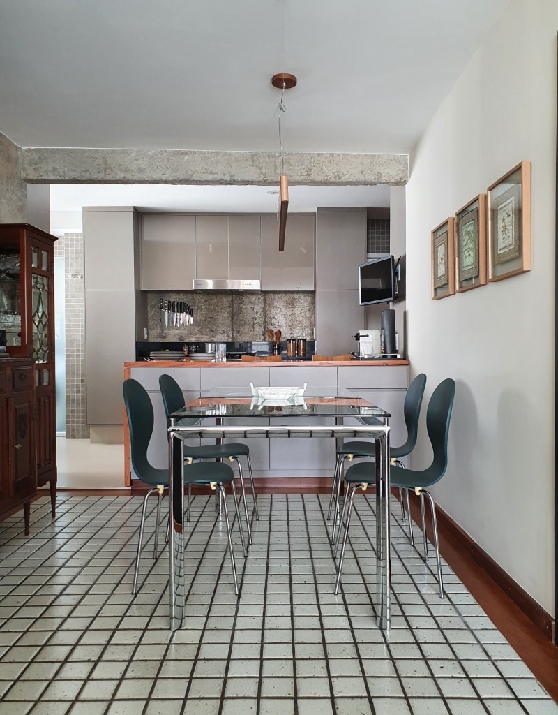 Sala de jantar com mesa de vidros e 4 cadeiras cinzas, cerâmica branca no piso, cristaleira de madeira com parede de fundo de cimento, com vista para cozinha, com armários cinzas e bancada amadeirada.