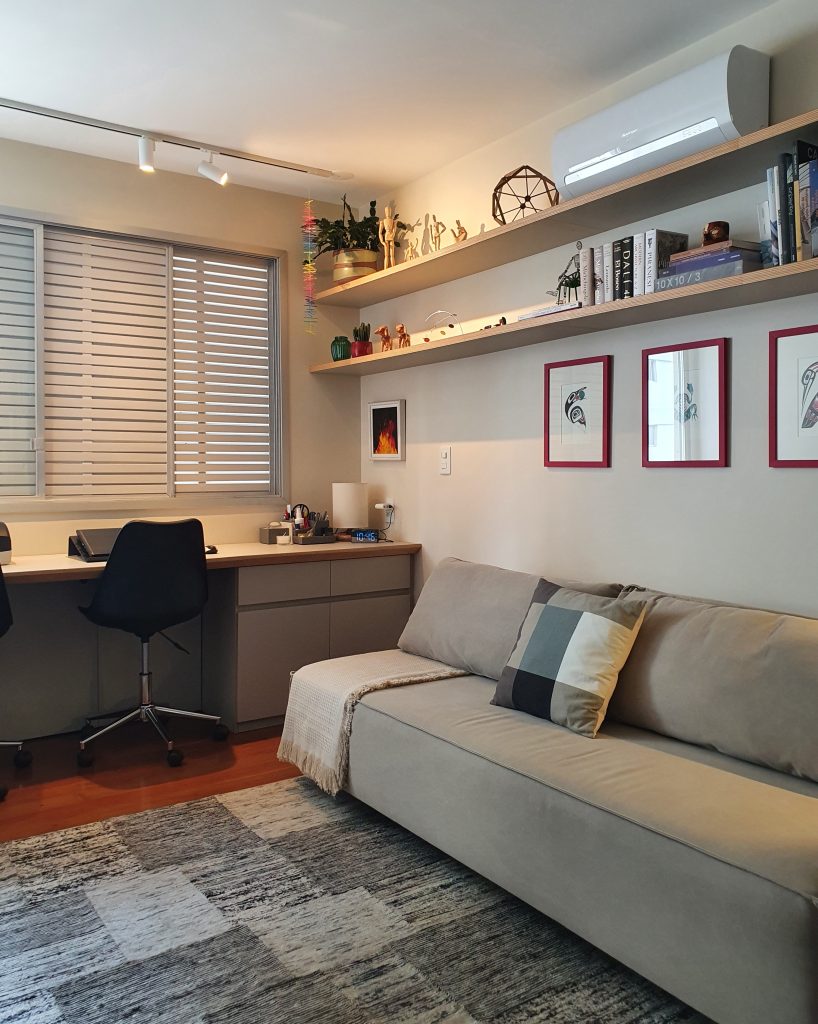 Sala de TV com piso de madeira, sofá cinza, espaço para home office com bancada extensa, prateleiras com livros e porta retratos.
