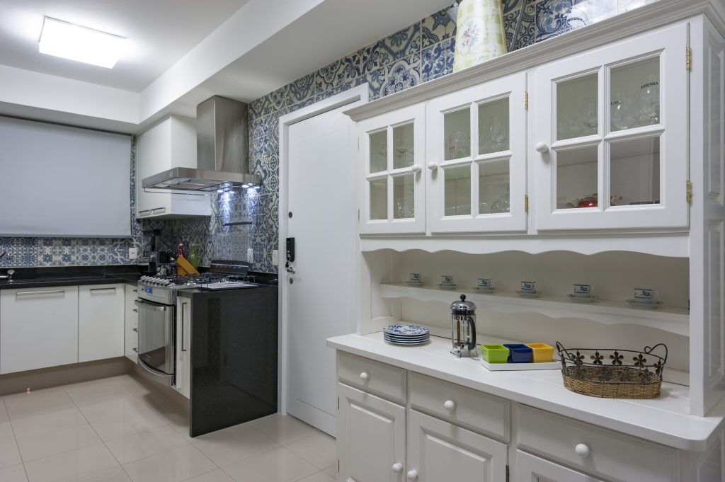 cozinhas com história e afeto -  Cozinha com azulejaria portuguesa, piso porcelanato branco, eletrodomésticos de inox e armários brancos clássico com portas de vidro e madeira trabalhada. 