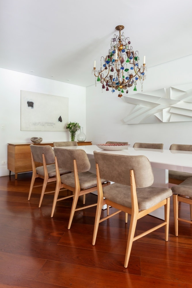 Sala de jantar com piso de madeira, mesa branca com cadeiras estofadas em tom neutro, móvel de madeira com vasos, paredes brancas e lustre colorido grande