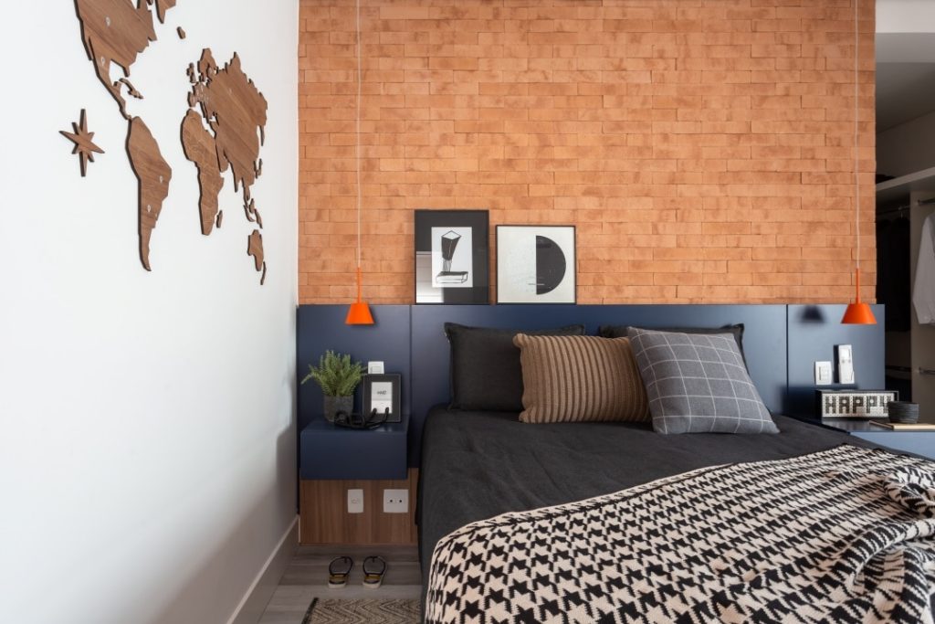Na parede lateral do dormitório, a reprodução do mapa mundi também remete às viagens do morador. No décor, o tijolinho aparente e, mais uma vez, o mix entre o azul presente na cabeceira de marcenaria e os pendentes laranjas
