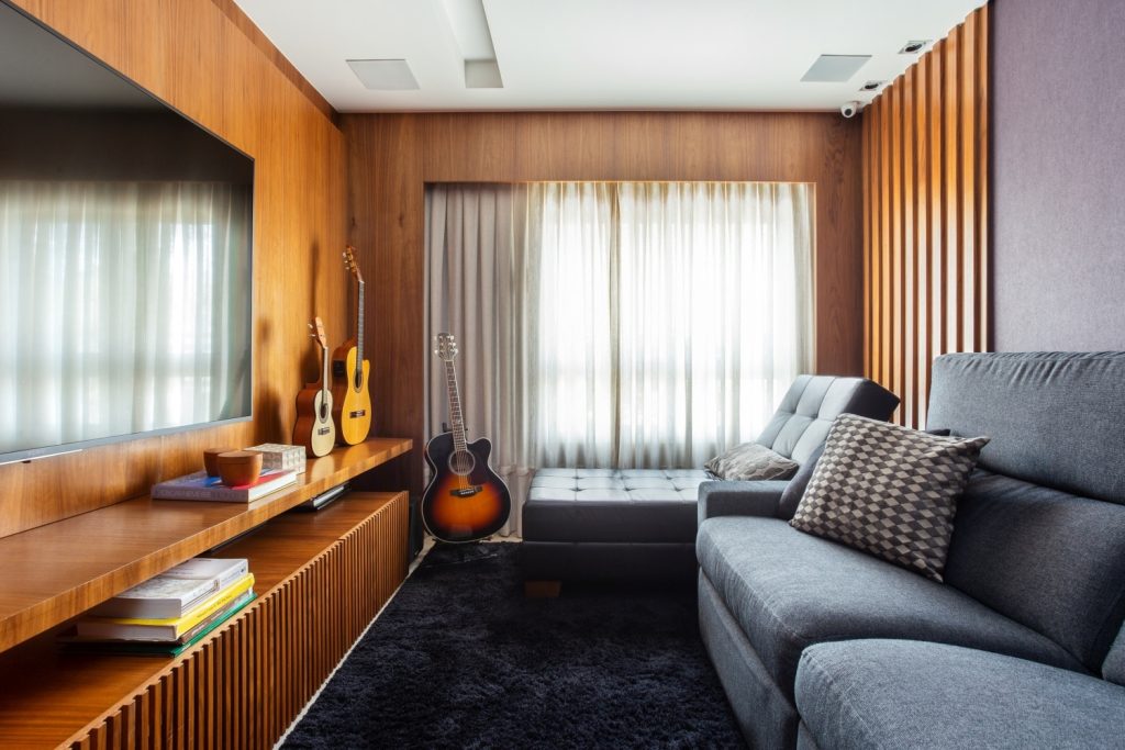 Sala com revestimento de madeira nas paredes, rack grande de madeira como painel de TV, sofá cinza e tapete cinza escuro