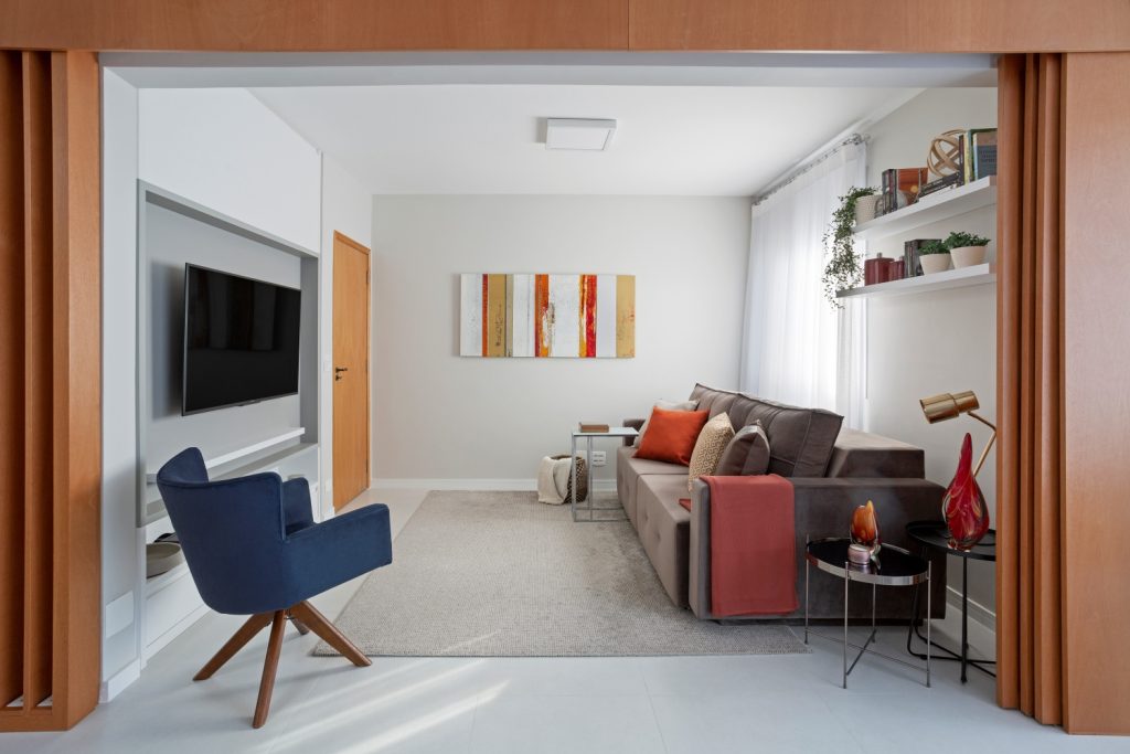 Sala com décor moderno, piso porcelanato branco, poltrona azul, sofá cinza, paredes brancas e objetos decorativos em tons vermelho e laranja