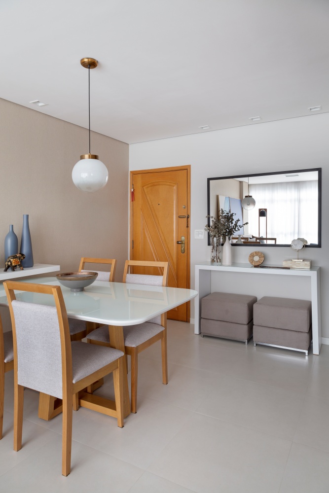 Sala de jantar com piso porcelanato branco, mesa de vidro branca com quatro cadeiras amadeiradas de estofado em tom neutro integrada a sala de estar