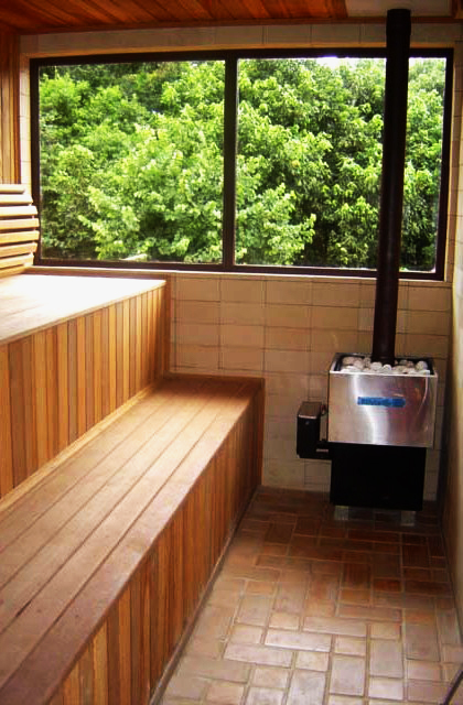 Na casa de campo, a sauna seca foi pensada para ter uma integração com a vegetação externa. Dessa forma, o ambiente recebeu vidros temperados nas janelas, placas refratárias nas paredes e área do banco e teto em madeira 