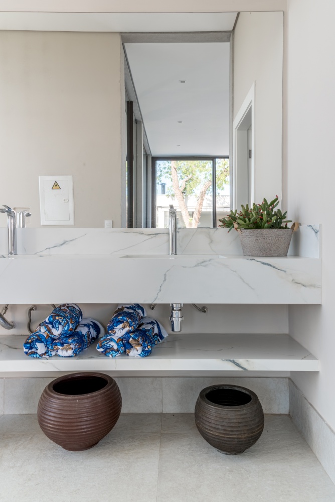 Banheiro com ducha para utilização pós sauna , com pia revestida de porcelanato marmorizado branco, piso em tom neutro e vasos decorativos no chão