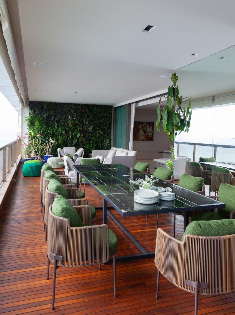 Sacada com mesa retangular grande verde e cadeiras amadeiradas com estofado verde, piso de madeira, no fundo jardim vertical com sofás branco e vasos de plantas decorativos.