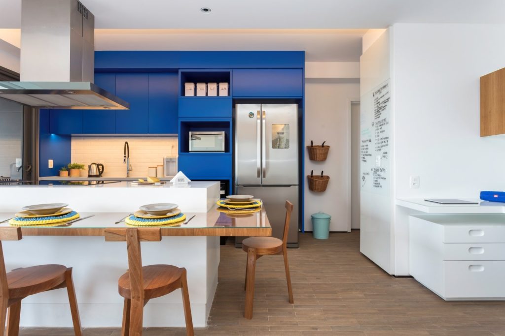 cozinha com ilha e bancada branca com três cadeiras de madeira, coifa de inox, piso de madeira, paredes brancas e armário azul