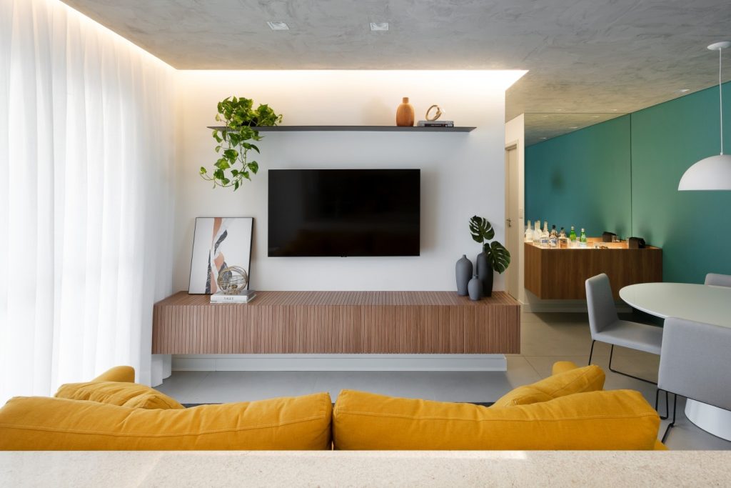 Sala de estar com sofá mostarda, rack amadeirada, TV na parede e prateleira com objetos decorativos