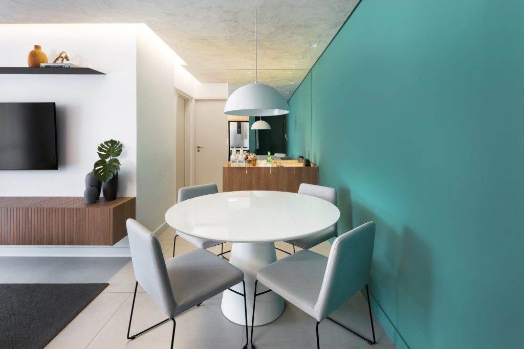 Canto da sala de jantar com parede verde agua, mesa redonda branca com quatro cadeiras e luminária pendente branca