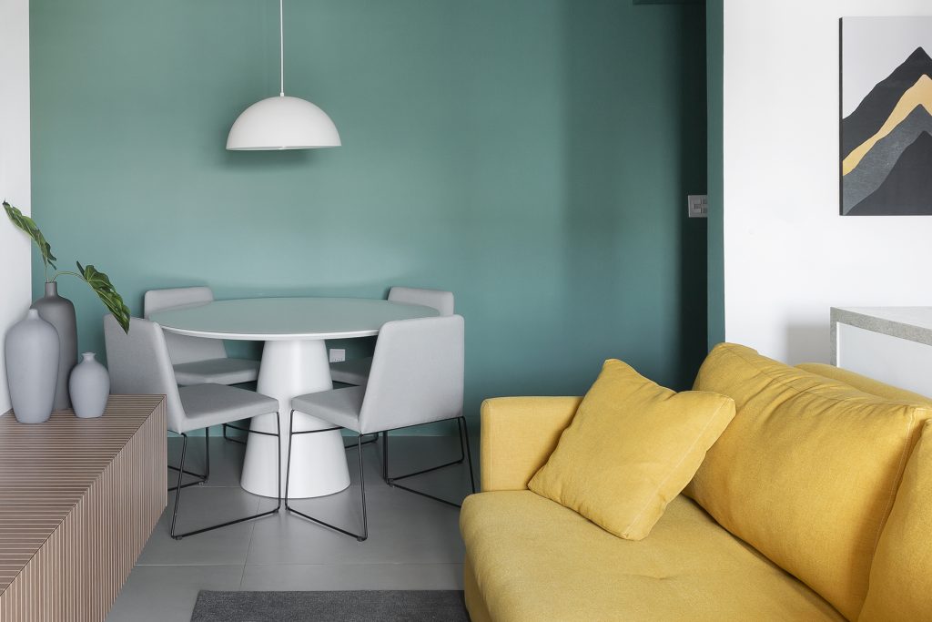 Necessidades do morar e trabalhar: Sala  de estar integrada a sala de jantar com pintura verde agua, sofá mostarda, rack amadeirada, mesa redonda branca com quatro cadeiras e piso cinza 