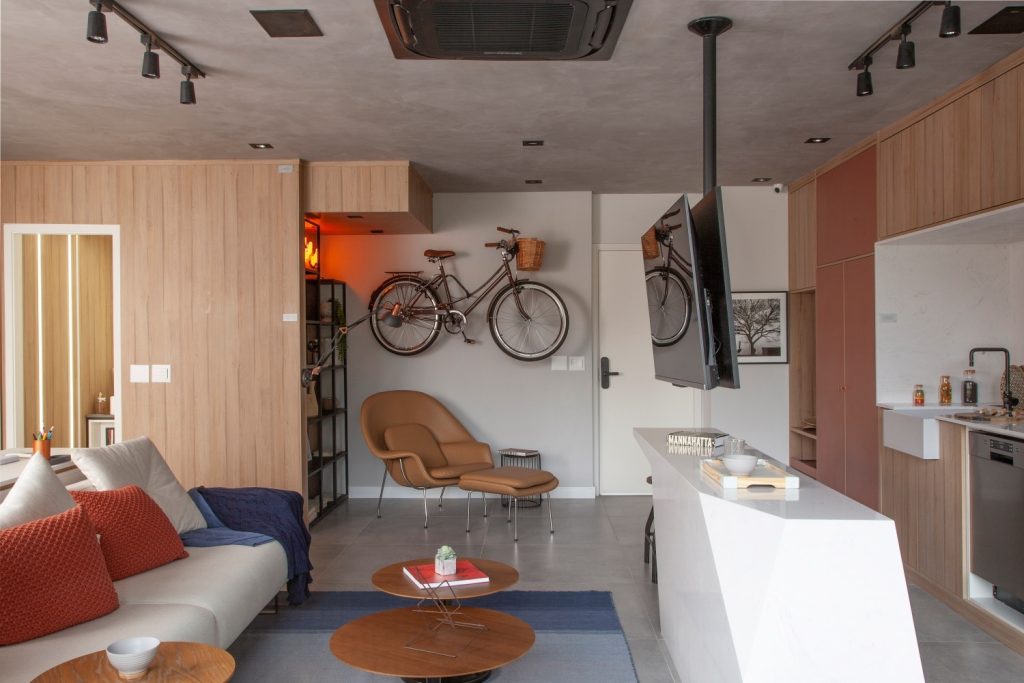 Reforma rápida: Sala de estar integrado a sala de jantar com sofá branco, e TV suspensa no teto, bancada branca, armários marrom e laranja com piso em tom neutro, no canto da parede um poltrona marrom e uma bicicleta pendurada na parede