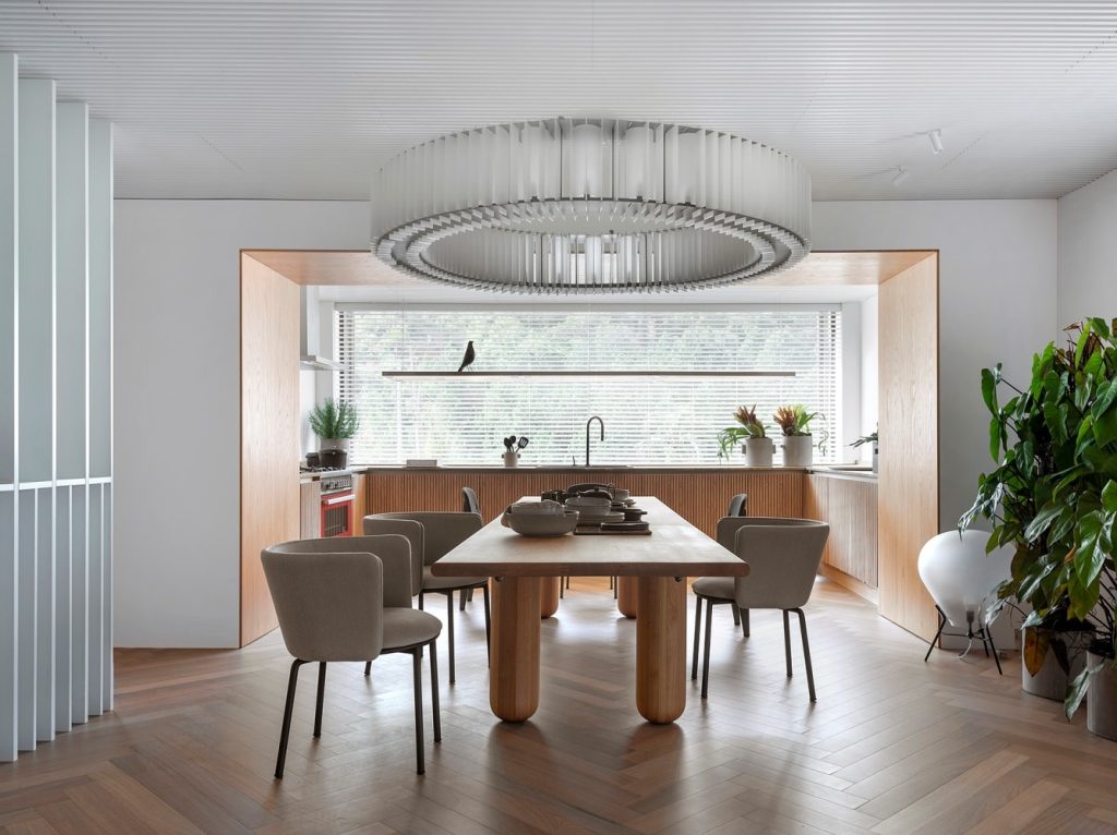 Cozinha com piso de madeira, mesa comprida de madeira com cadeiras estofadas em tom neutro, armários de madeira e luminária grande redonda sob a mesa.