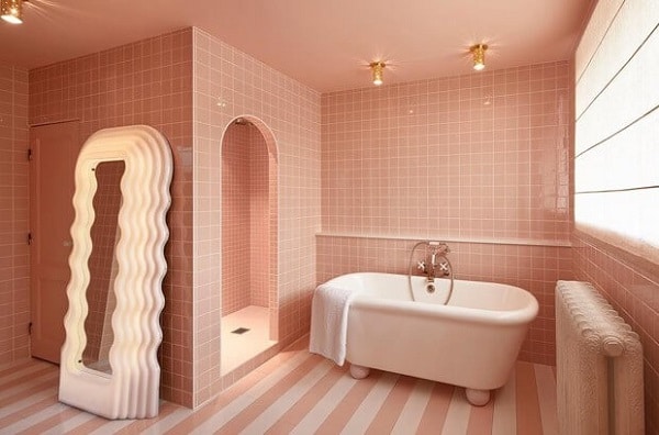 Banheiro com cerâmica rosa na parede, banheira branca, piso listrado rosa e branco, espelho com moldura ondulado branco
