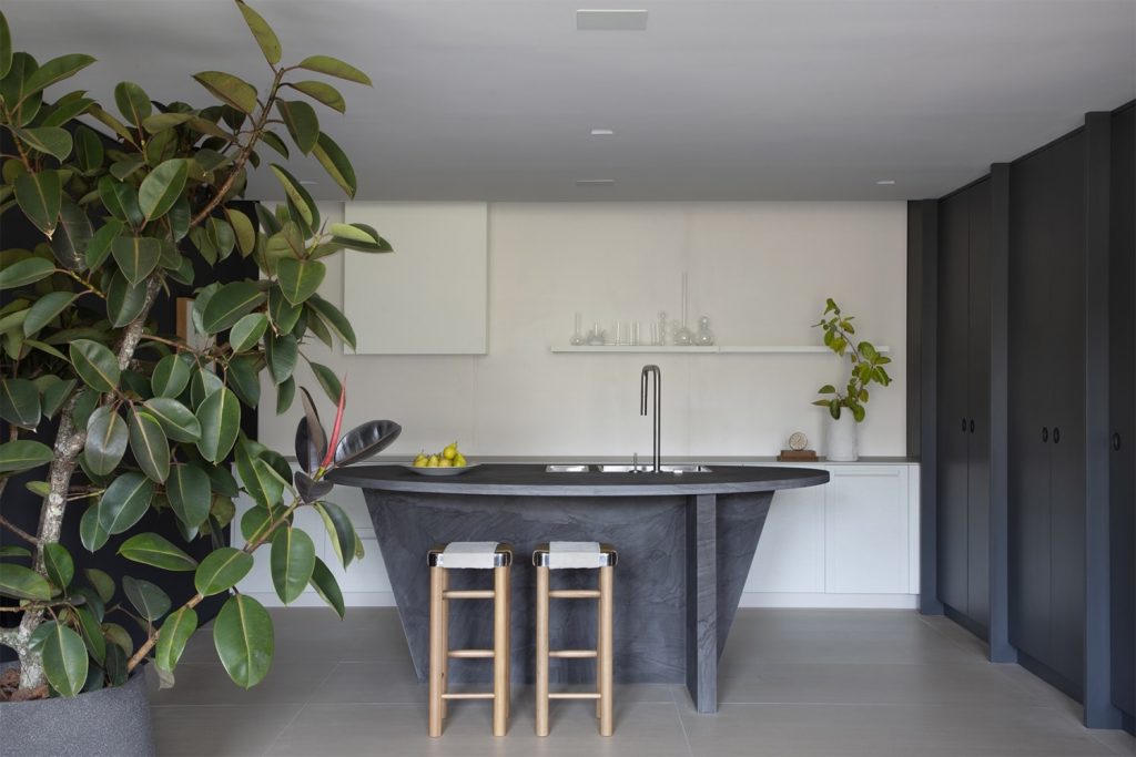 Cozinha com extensão de living, com ilha central no formato orgânico em cimento queimado, piso cinza, duas baquetas d emadeira, armários pretos e parede branca