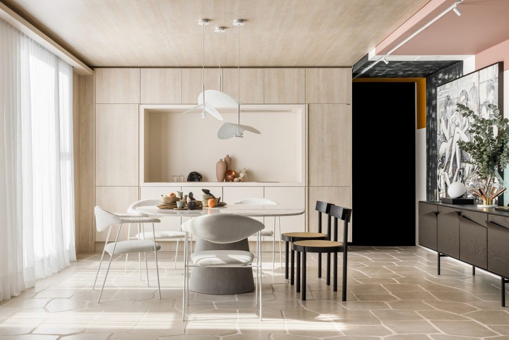 Cozinha com piso em tom neutro, mesa redonda branca com cadeiras brancas, armário em tom claro, luminária pendente branca e quadros decorativos