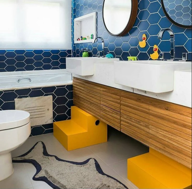 Banheiro infantil com parede e banheira revestidas com azulejo hexagonal, bancada branca com duas pia embutida, armário de madeira, piso porcelanato branco com degrauzinhos removível amarelo.